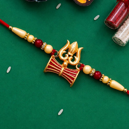A gold Trishul and Damaru rakhi with red and cream beads on a red thread, against a green background.