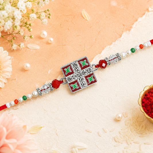 A red thread rakhi with a square silver charm featuring red and green enamel patterns, surrounded by white flowers and