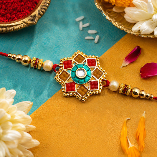 A gold-plated rakhi with a central teal and red floral design, surrounded by rice grains and red powder.
