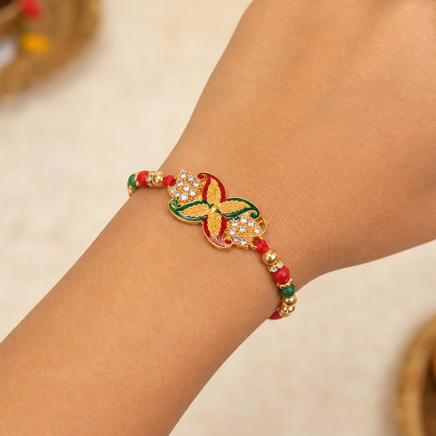 A person's arm wears a red thread bracelet with a golden, red, and green flower motif, featuring clear stones and small red,