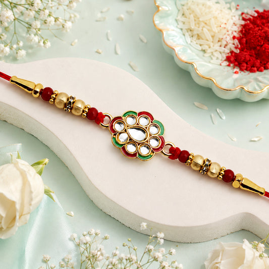 A red thread rakhi with a central kundan floral design and red and gold beads, surrounded by white flowers and a bowl of