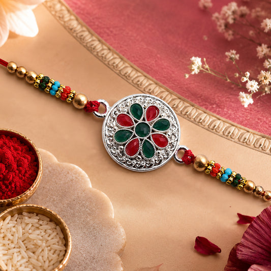 Silver-plated floral rakhi with red and green stones, gold beads, and red thread, next to kumkum and rice