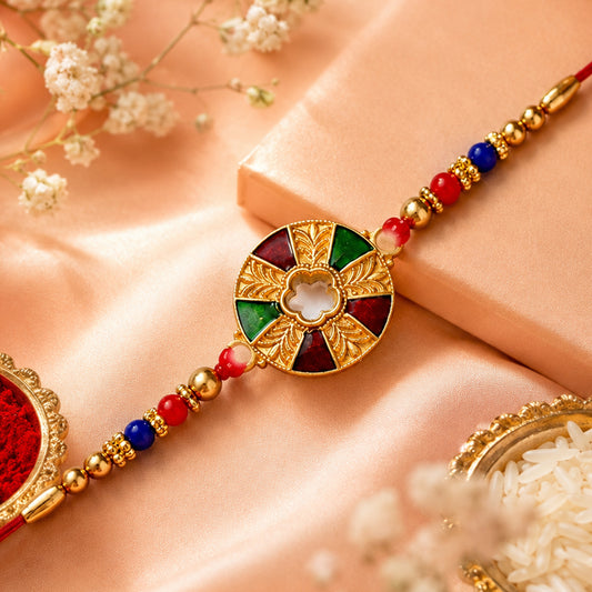 A gold Kundan Rakhi with red, green, and maroon enamel, on a red beaded thread, against a light pink background.