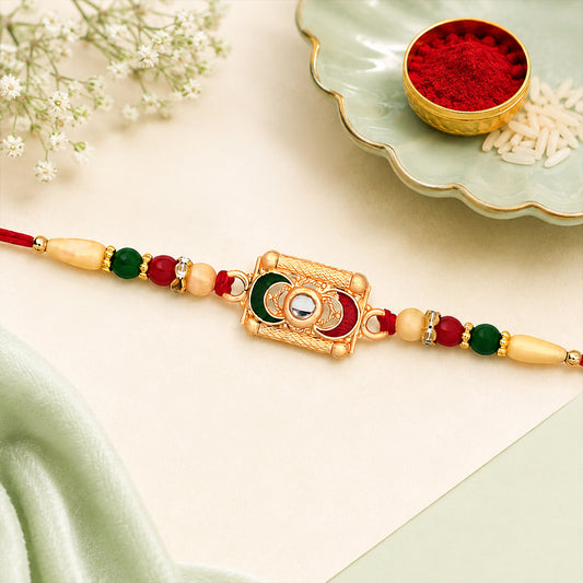A gold-plated Meenakari rakhi with red thread, green and red beads, and a central clear stone, beside a small bowl of red