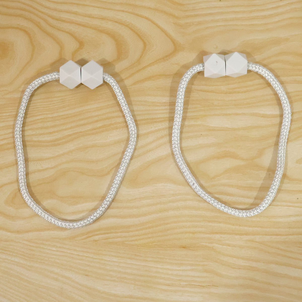 Two white braided rope curtain tiebacks with geometric white plastic fasteners laid flat on a light wooden surface.