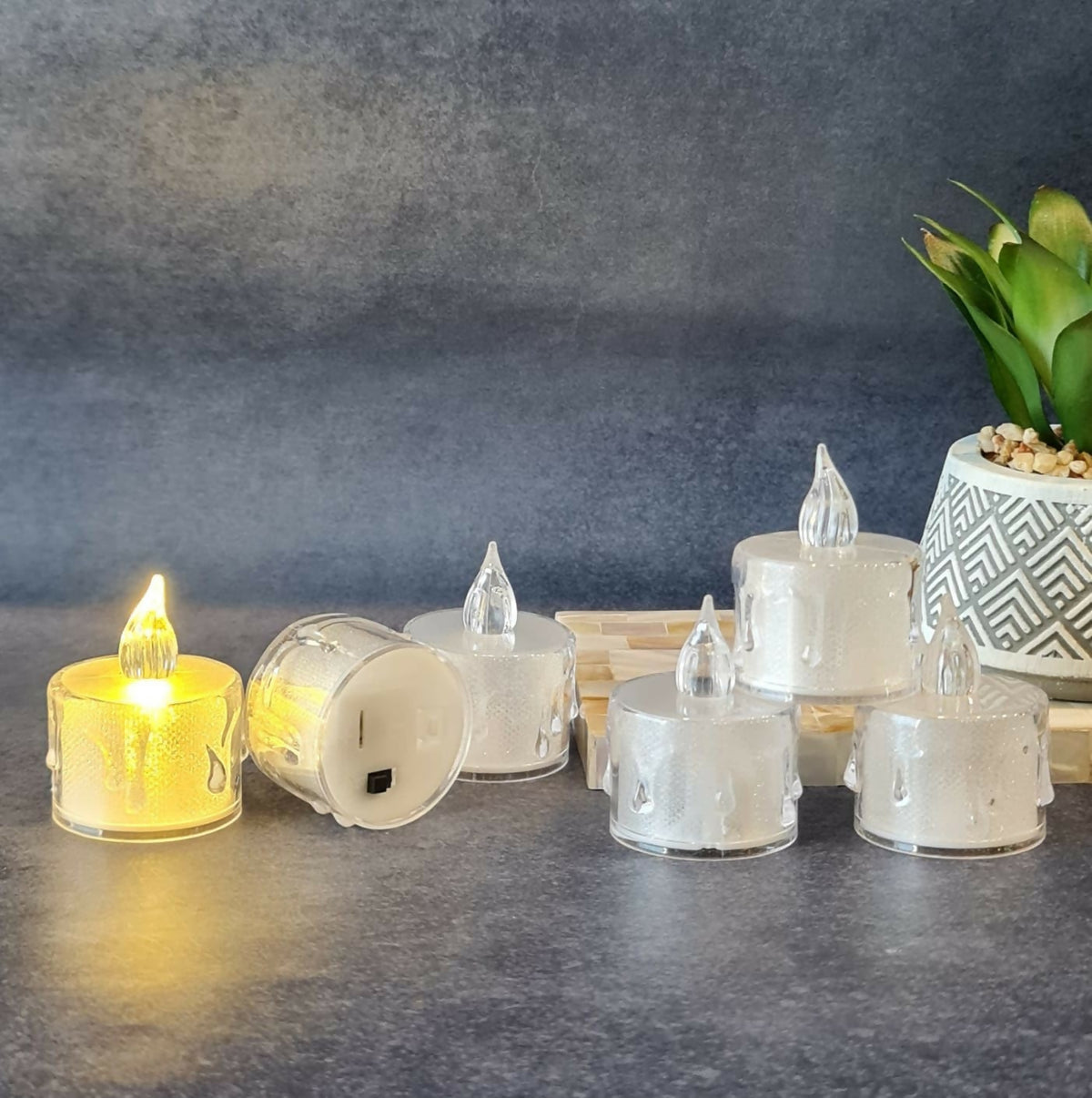 Six clear plastic LED candles, one lit, arranged on a dark grey surface next to a potted green plant.