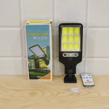 The black solar LED street light, its remote, and its product box are displayed on a wooden surface against a white tiled