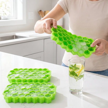 A person bends a green silicone tray, releasing small hexagonal ice cubes into a glass of water with mint and lemon. Two
