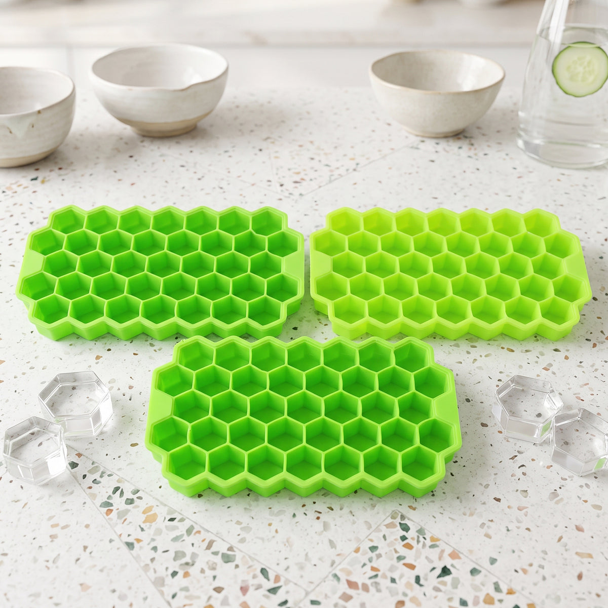 Three bright green silicone ice cube trays with hexagonal compartments sit on a speckled white surface, with two clear