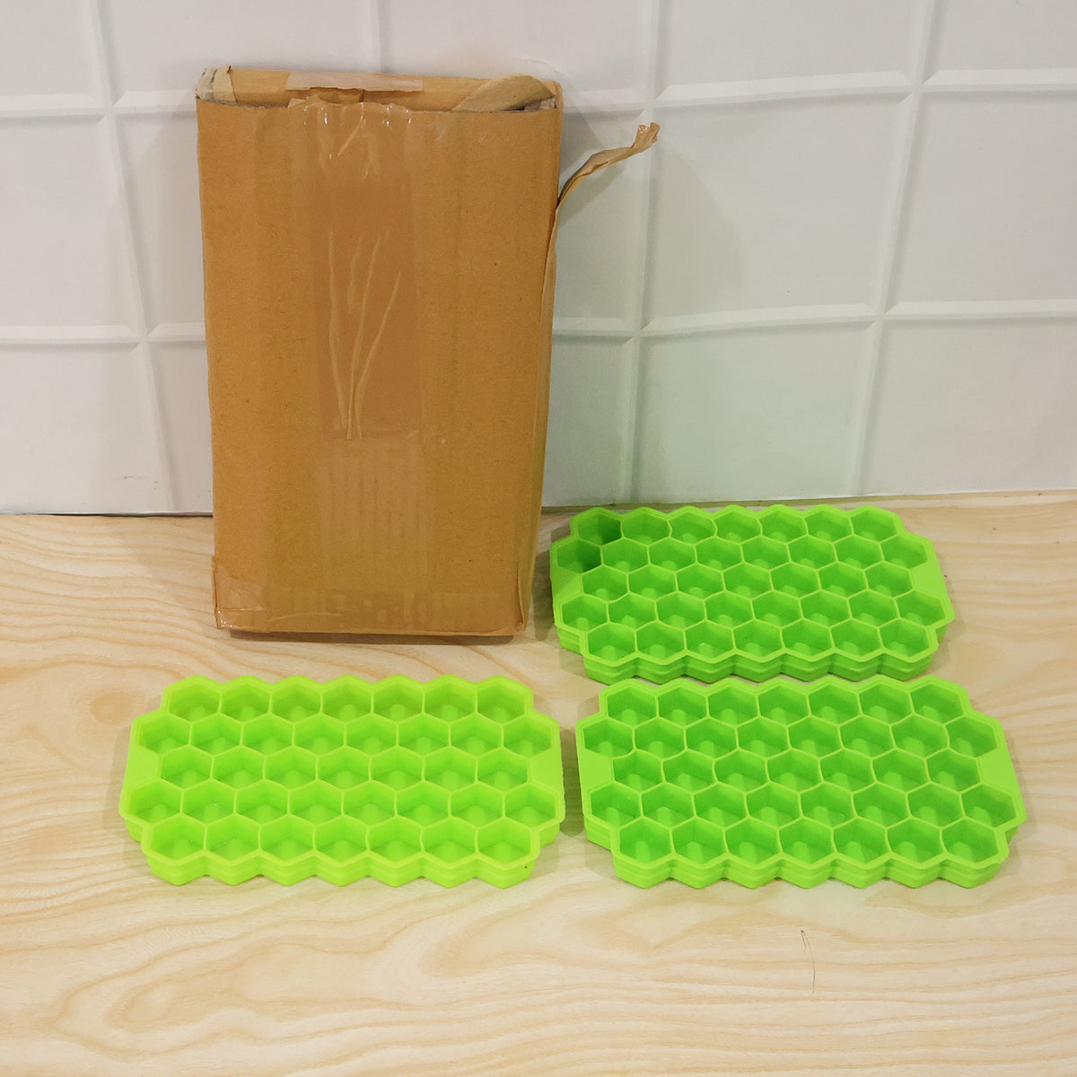 Three green silicone honeycomb ice cube trays are placed on a light wooden floor next to a tall, narrow cardboard box.
