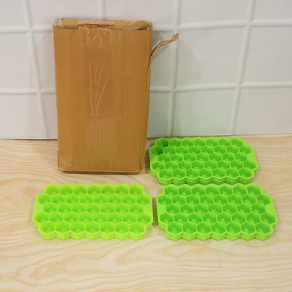 Three green silicone honeycomb ice cube trays are placed on a light wooden floor next to a tall, narrow cardboard box.