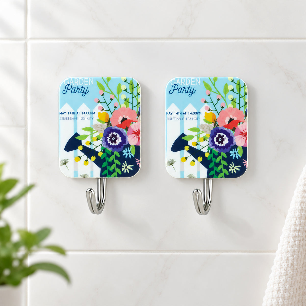 Close-up of two garden party themed adhesive hooks on a white tiled wall, featuring colorful floral designs.