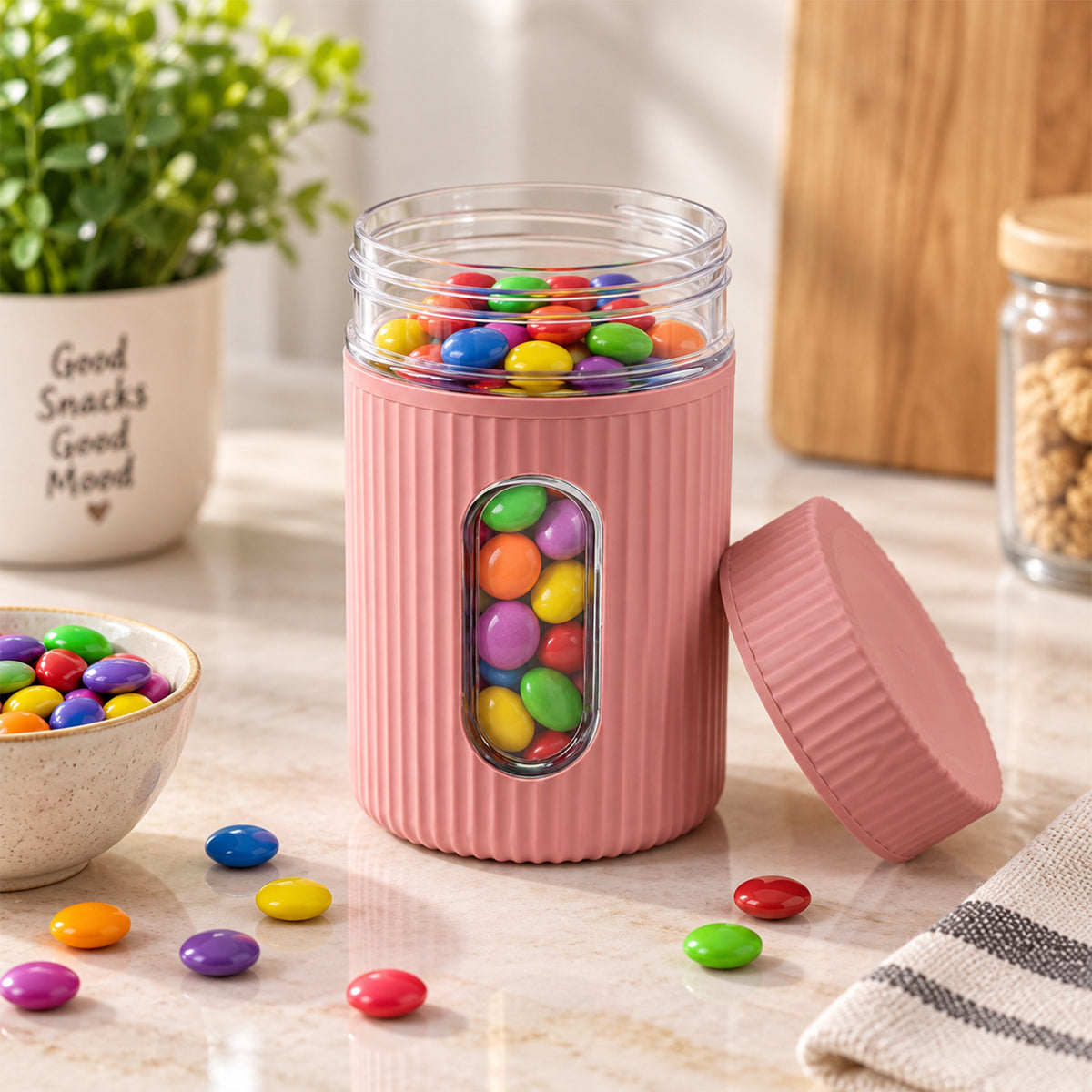 A pink ribbed storage jar with a clear window filled with colorful candies, its lid rests beside it on a marble counter.