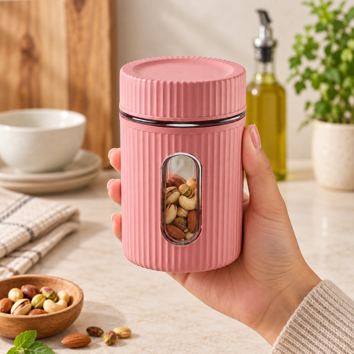 A hand holds a pink ribbed storage jar with a clear window filled with mixed nuts, on a kitchen counter.