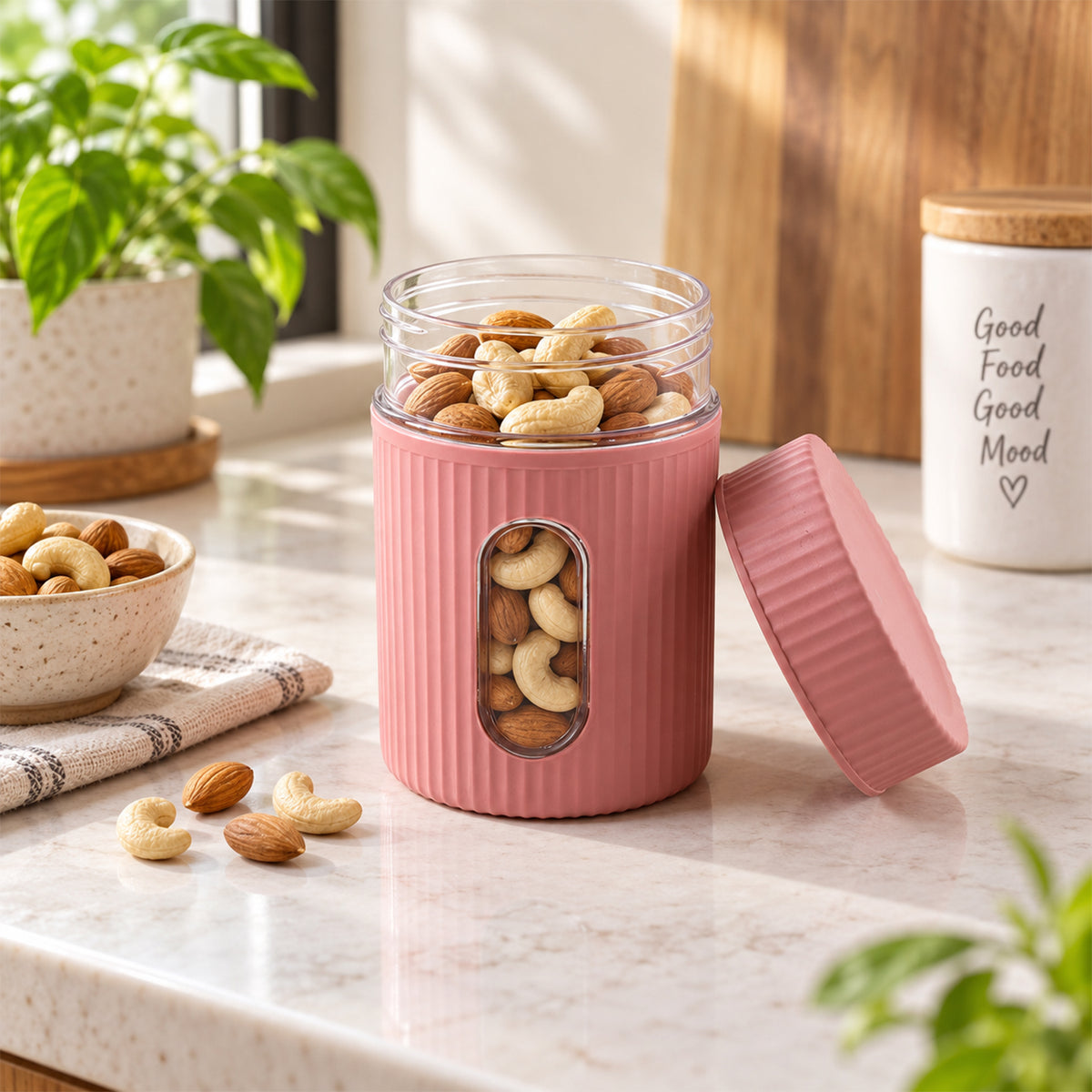 A pink ribbed storage jar with a clear window filled with almonds and cashews, its lid rests beside it on a counter.