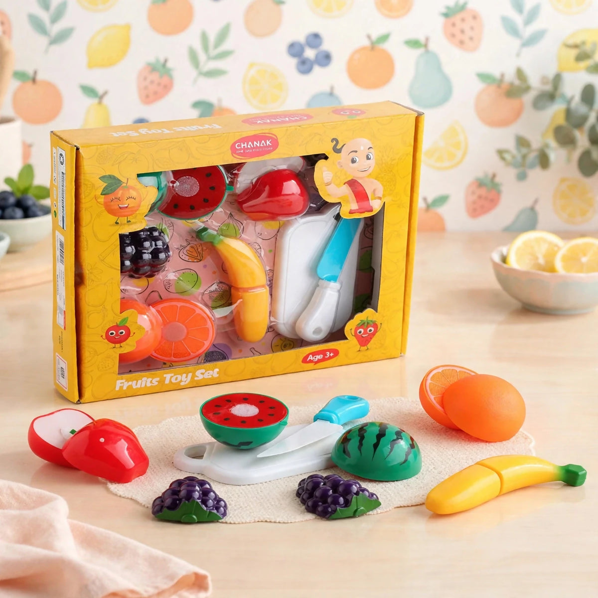 A yellow boxed set of plastic fruit toys, a knife, and a cutting board, with some pieces arranged on a white mat.