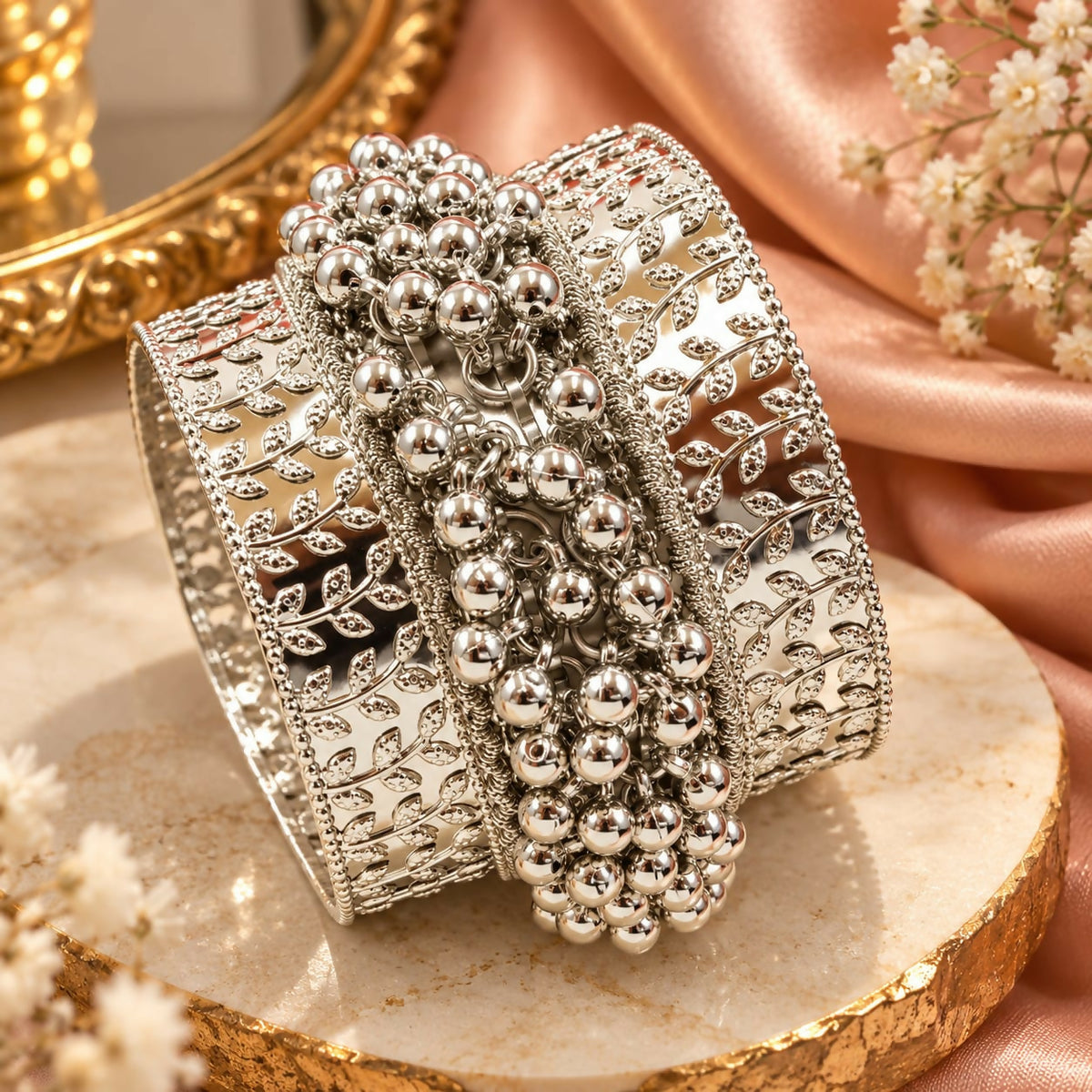 A wide silver-tone bangle with leaf patterns and a central band of dangling ghungroo beads rests on a marble surface.