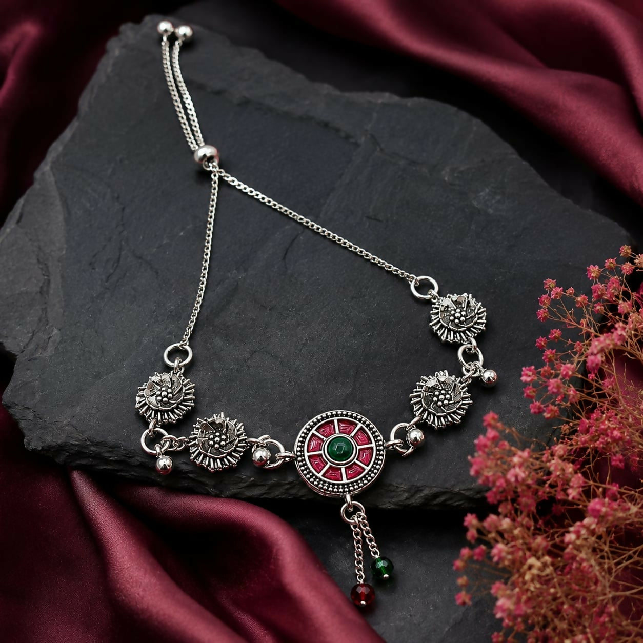 The oxidized silver bracelet with a red and green enamel mandala center rests on a dark slate surface with a burgundy cloth.