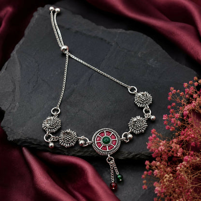 The oxidized silver bracelet with a red and green enamel mandala center rests on a dark slate surface with a burgundy cloth.