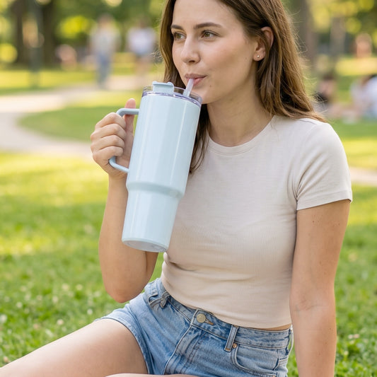 A woman sits on grass, sipping from a large light blue handled tumbler with a straw.