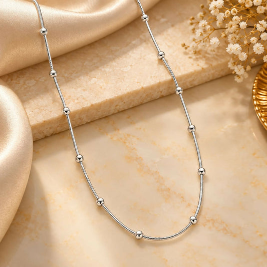 A silver-toned snake chain necklace with evenly spaced round beads rests on a light beige fabric.