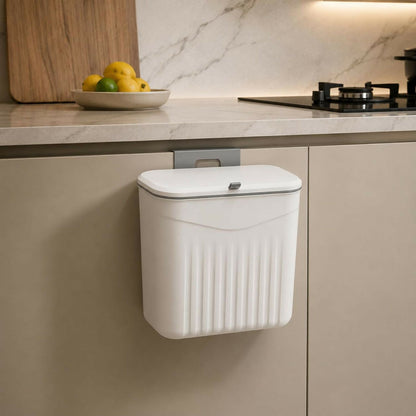 A white plastic dustbin with a lid hangs from a grey hook on a light brown kitchen cabinet, with a bowl of fruit nearby.