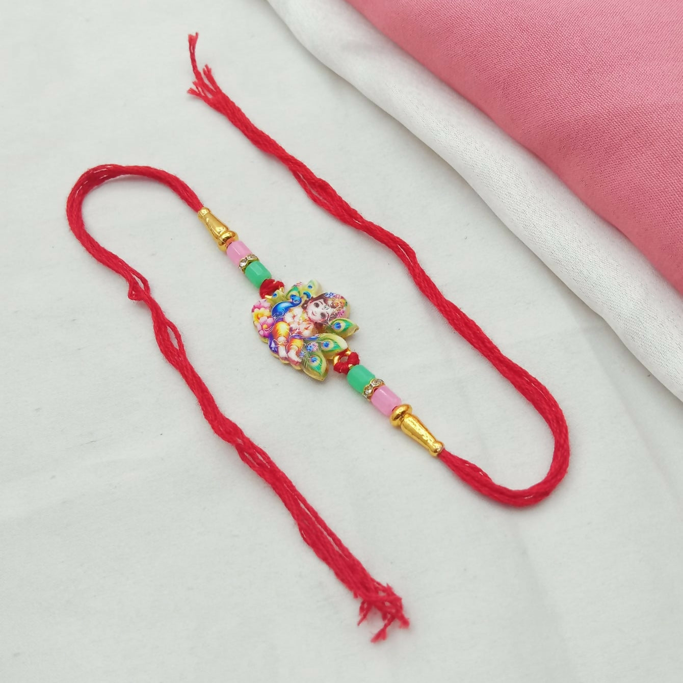 A red thread Rakhi with a Krishna and peacock feather charm lies on a white and pink fabric background.