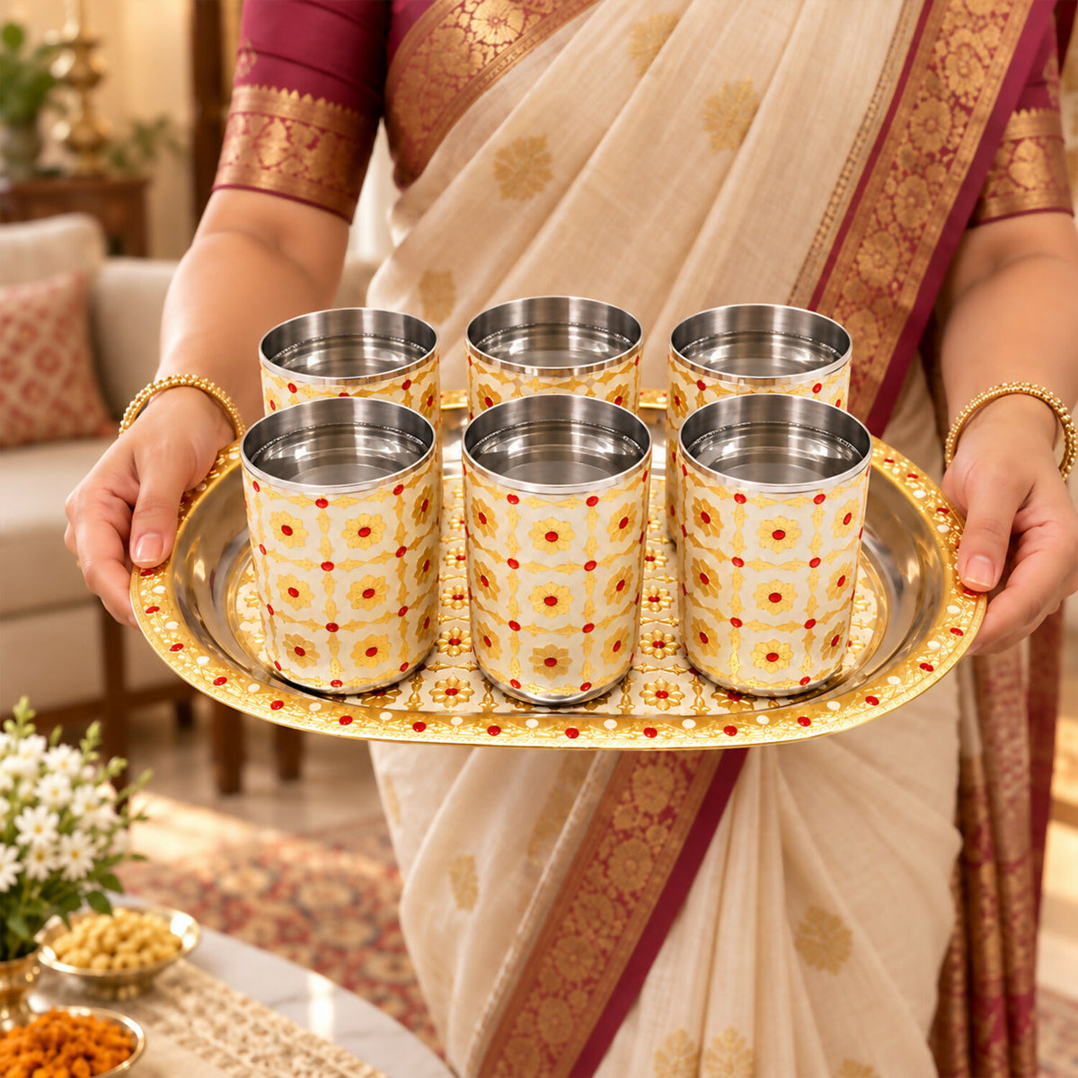 A person in traditional attire holding an oval serving tray with six stainless steel glasses filled with water.