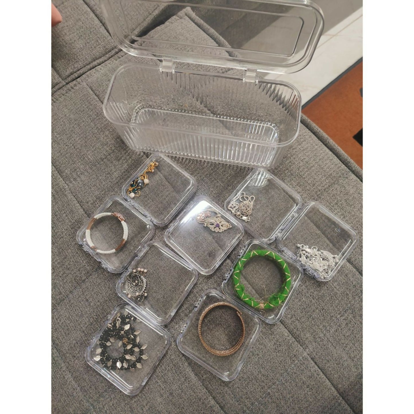 A clear, ribbed plastic storage box with an open lid is on a gray fabric surface, surrounded by nine small square containers