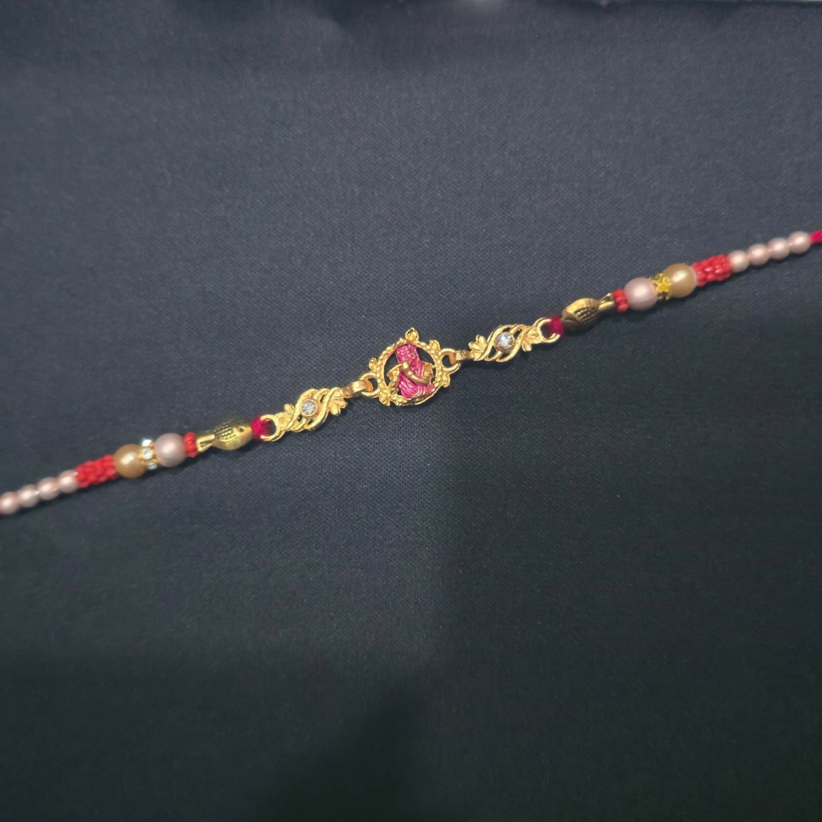 A gold and pink Lord Krishna Rakhi with red and white beads is centered on a plain black background.
