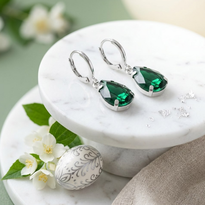 Green teardrop crystal earrings with silver hooks on a white marble surface, next to a decorative egg and flowers.
