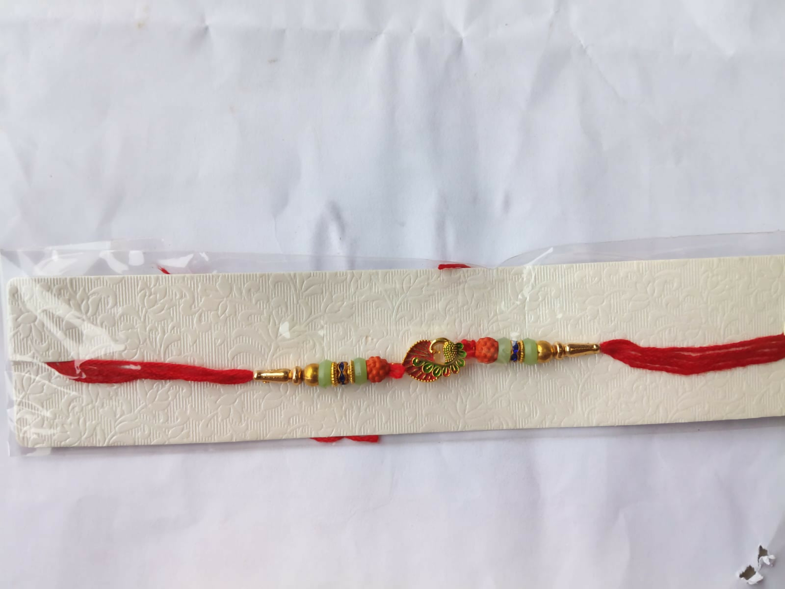 A red and gold bracelet with a peacock charm, orange and light green beads, and blue accents, packaged in clear plastic on