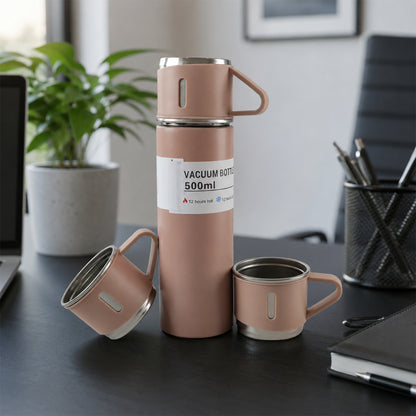 A pink vacuum flask and two matching cups are displayed on a dark office desk with a laptop, plant, and pen holder.