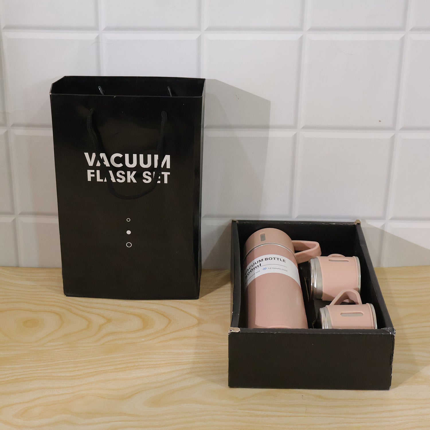 A black gift bag and a black box containing a pink vacuum flask set are placed on a wooden surface against a white tiled