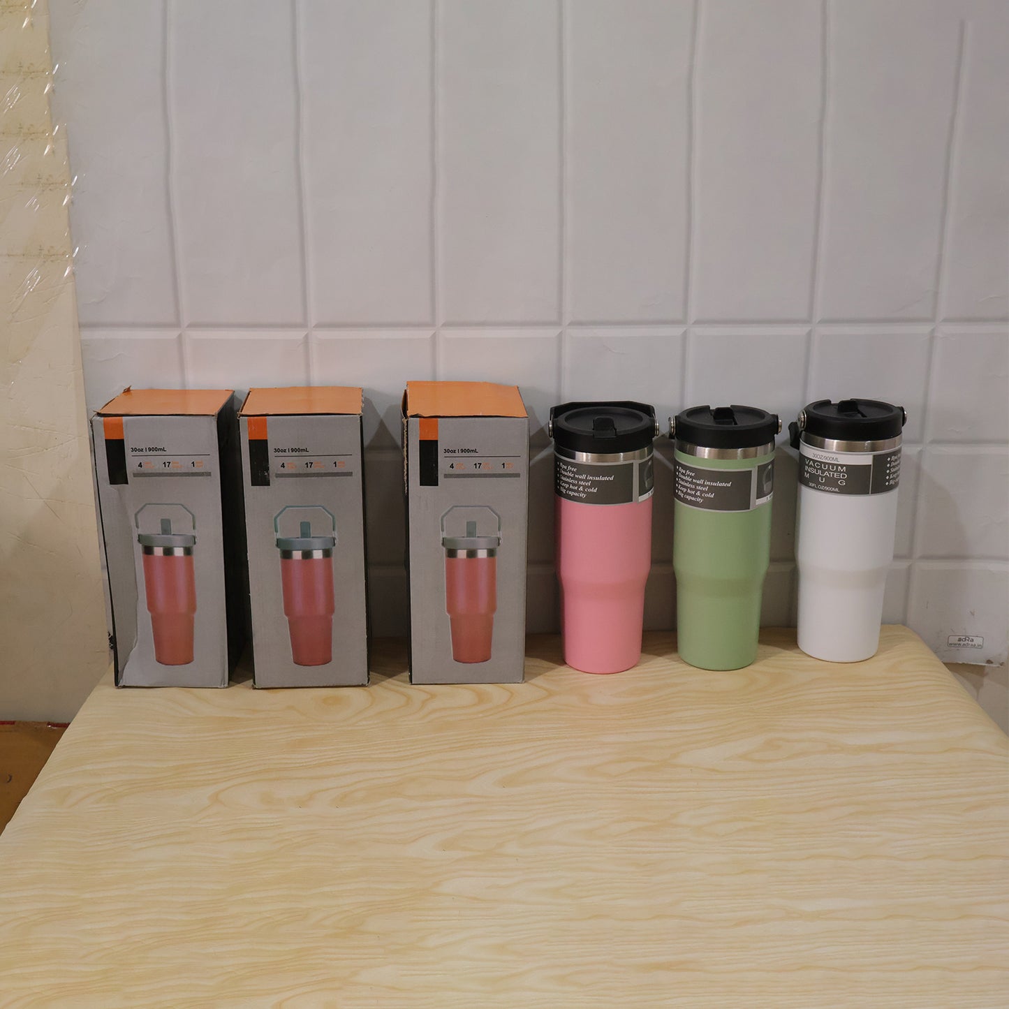 Three boxed pink tumblers and three unboxed tumblers in pink, green, and white sit on a wooden table.