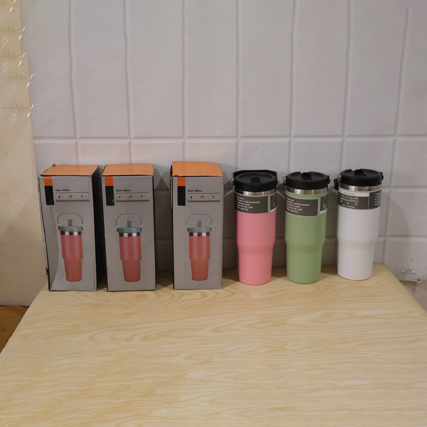 Three boxed pink tumblers and three unboxed tumblers in pink, green, and white sit on a wooden table.
