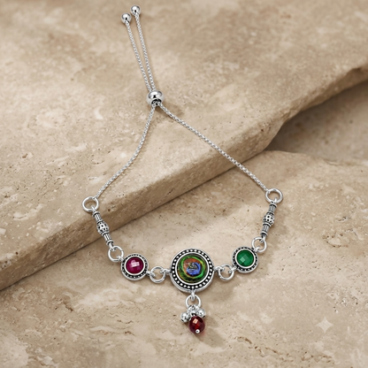 The antique silver adjustable bracelet with a peacock eye cabochon and maroon and green side stones rests on a beige stone