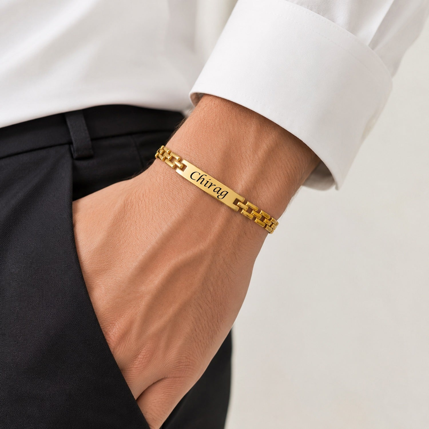 A man's hand, wearing a white shirt and black pants, displays a gold-plated wristband with "Chirag" engraved on its central
