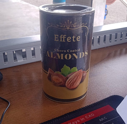 The Effete Choco Coated Almonds can is on a wooden desk with a black mouse pad and office items.