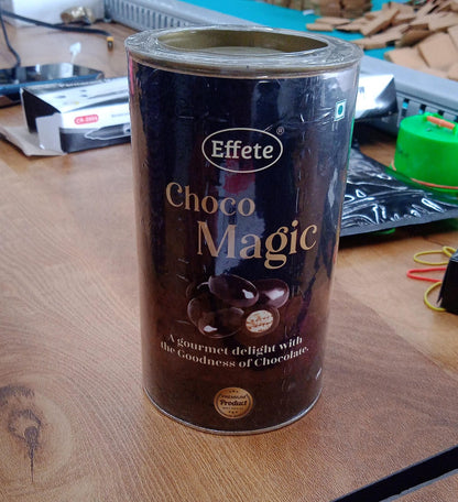 A brown cylindrical tin of "Choco Magic" candy sits on a wooden table in a casual indoor setting.