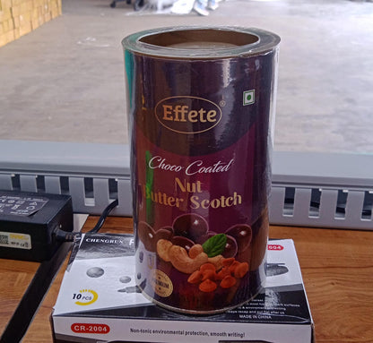 A purple and gold tin of chocolate-coated nuts sits on a white box on a wooden surface, with a black power adapter nearby.