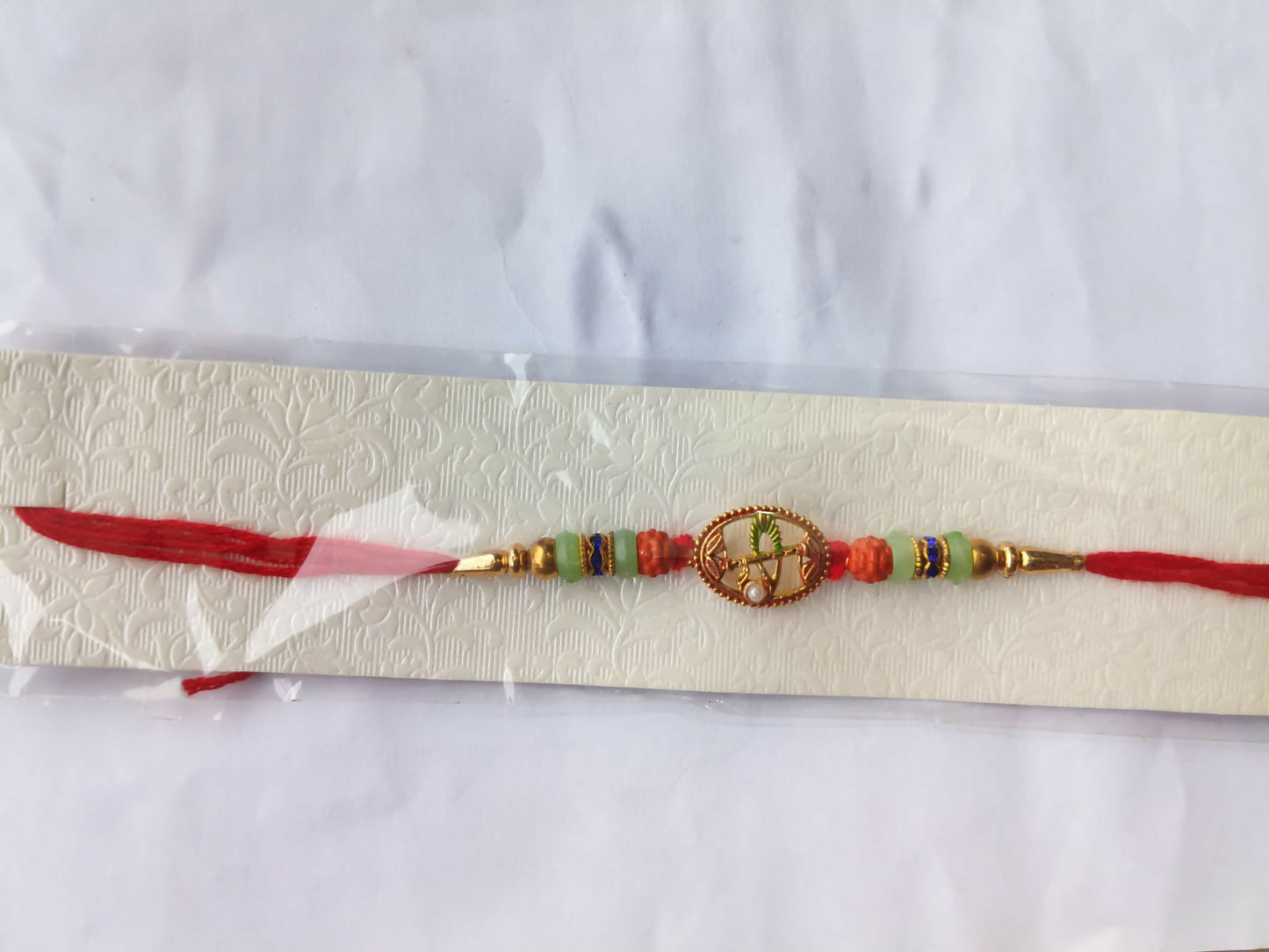 A red string bracelet with a central oval charm and colorful beads, packaged in clear plastic on a textured white card.