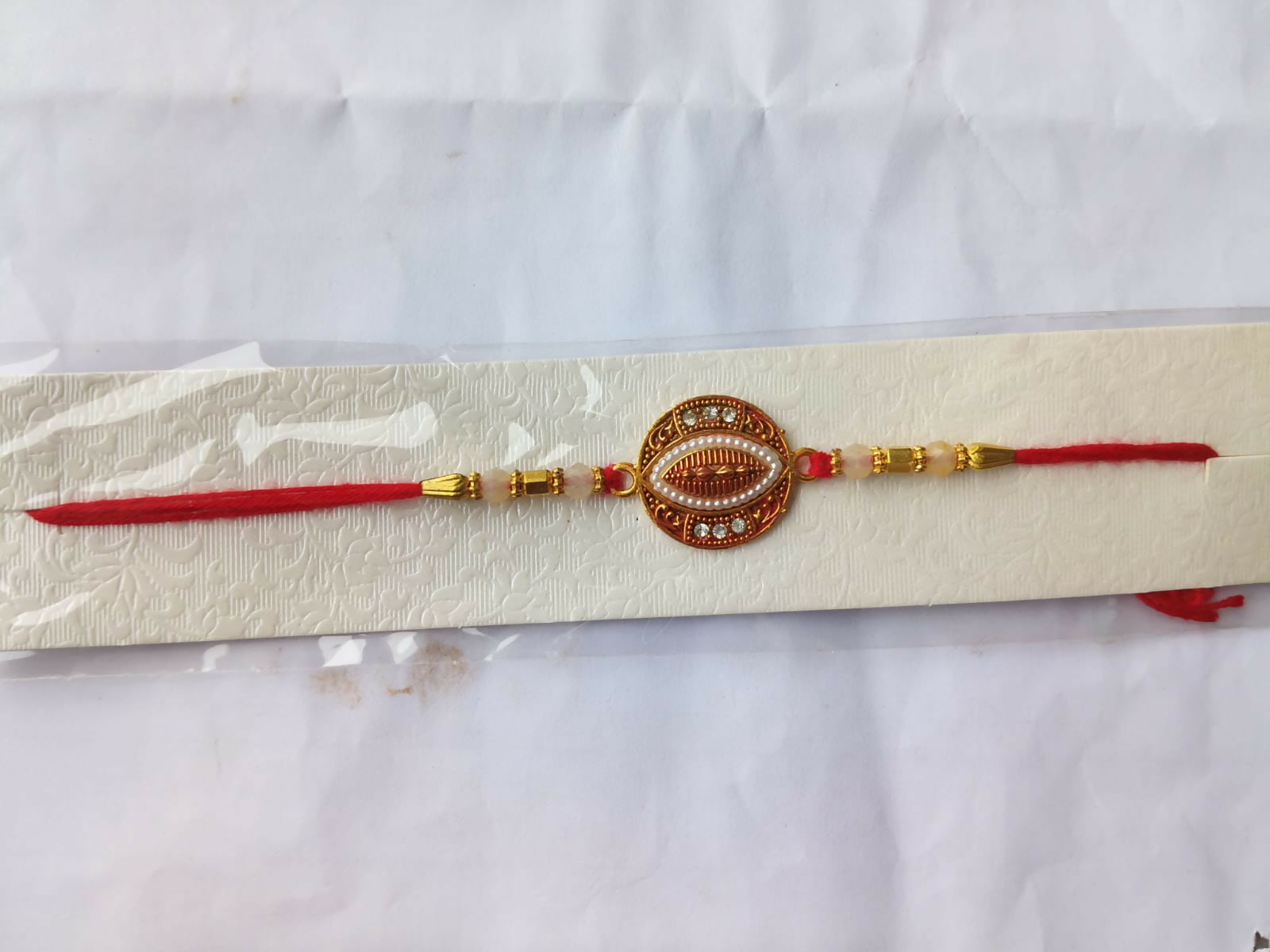 A red and gold bracelet with an oval centerpiece, pearls, and stones, is presented on a white textured card against a plain
