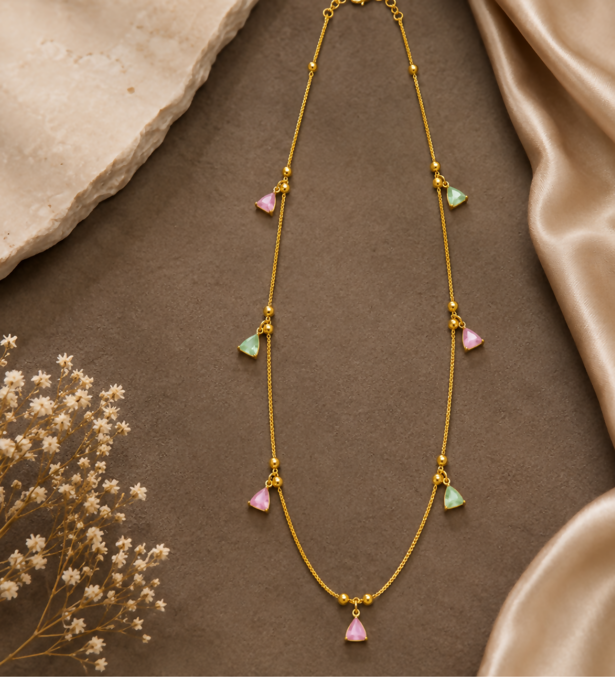 A gold-plated necklace with pink and mint green triangular charms is arranged on a brown surface with a silk cloth.