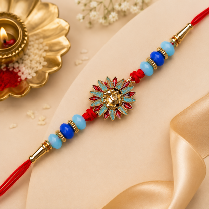 A red thread bracelet with a gold Ganesh charm, blue and light blue beads, and gold accents, next to a lit diya and rice.