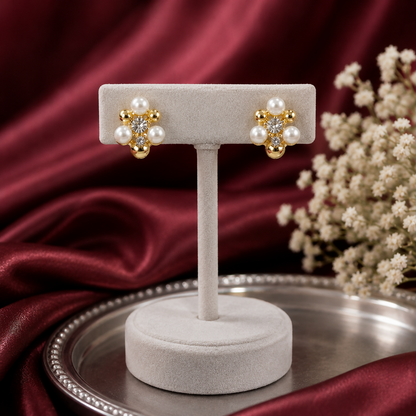 A pair of gold-plated floral stud earrings with crystals and mini pearls displayed on a light gray velvet earring stand.