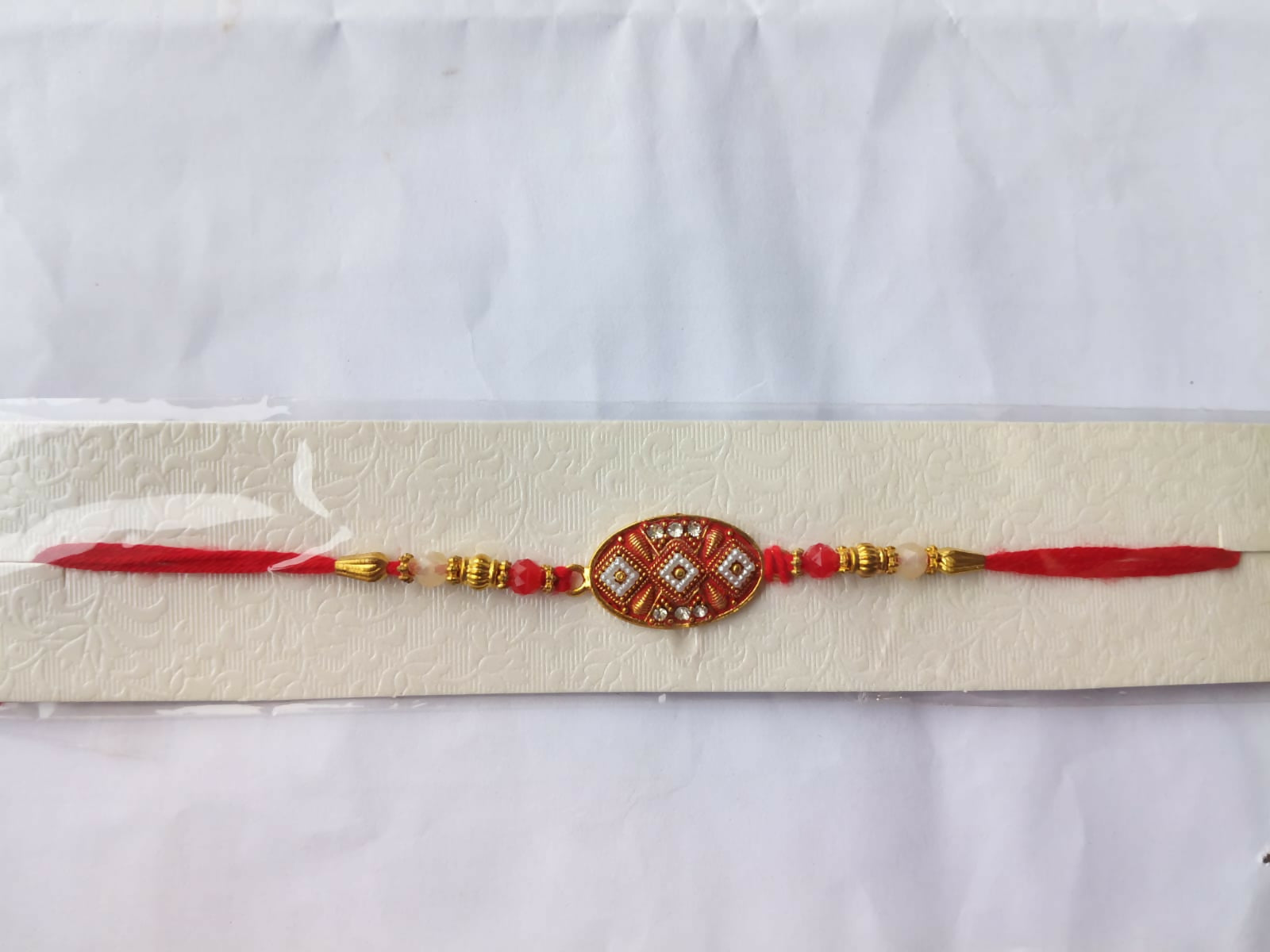 The red and gold Rakhi is packaged on a textured white card, sealed in clear plastic against a plain white background.