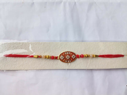 The red and gold Rakhi is packaged on a textured white card, sealed in clear plastic against a plain white background.