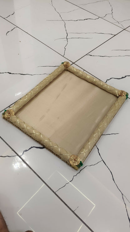 A simple beige fabric gift tray with gold braided edges and green corner accents, resting on a white tiled floor.