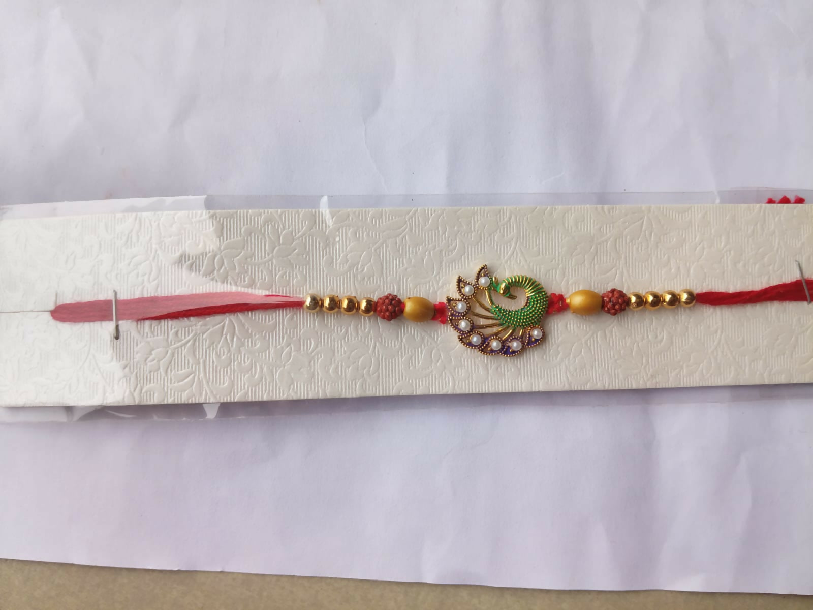A red thread with gold beads and a central green and purple peacock design with white pearls is displayed on a textured